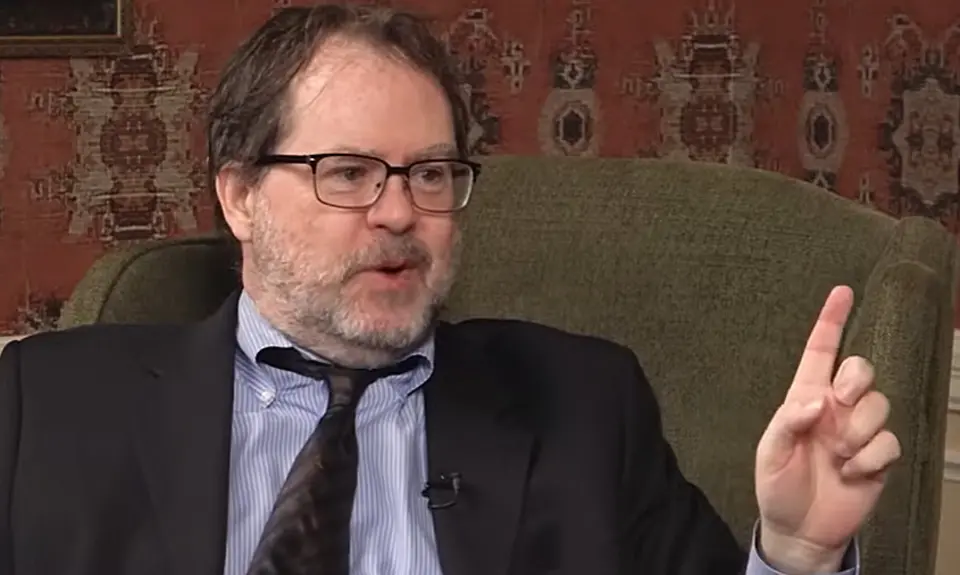 John Zmirak, a rumpled white guy sitting in an armchair, gestures with his hand while speaking with Eric Metaxas, who is off-camera. 