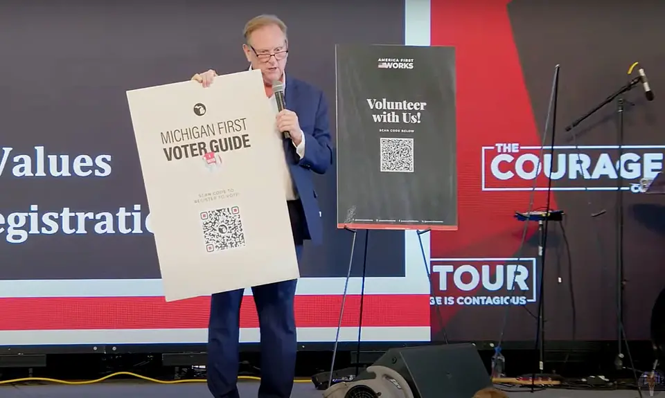 Richard Rogers stands on stage holding a placard with a QR code for voter registration. Logo of the Courage Tour is visible behind him. 