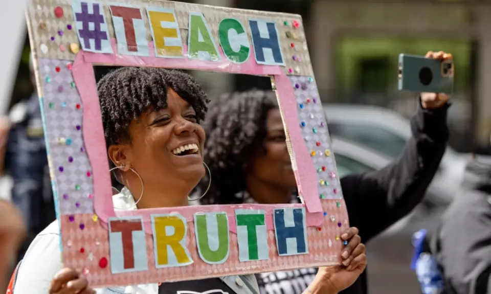 Woman holding a sign framing her face  that says #TeachTruth