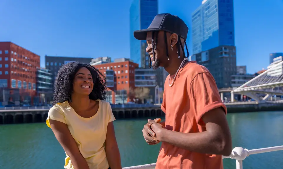 Photo of a young Black man and woman walking along a body of water in a city