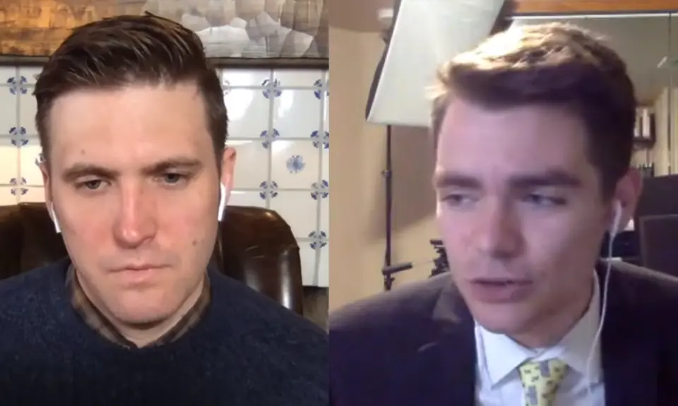 Nick Fuentes stands in front of American flag with a Trump campaign hat on