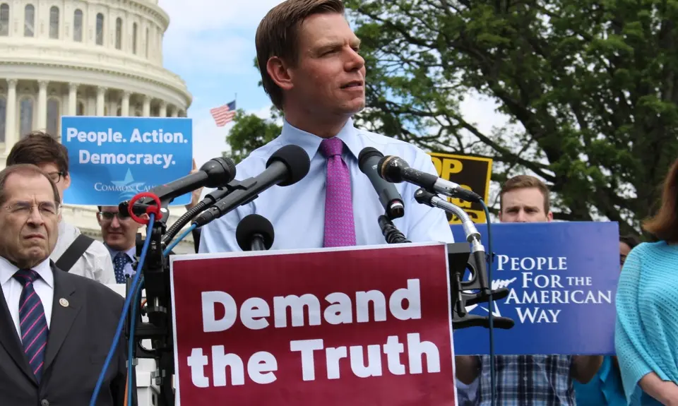 Man in tie speaking at podium in front of crowd. Podium has sign that says Demand the Truth.