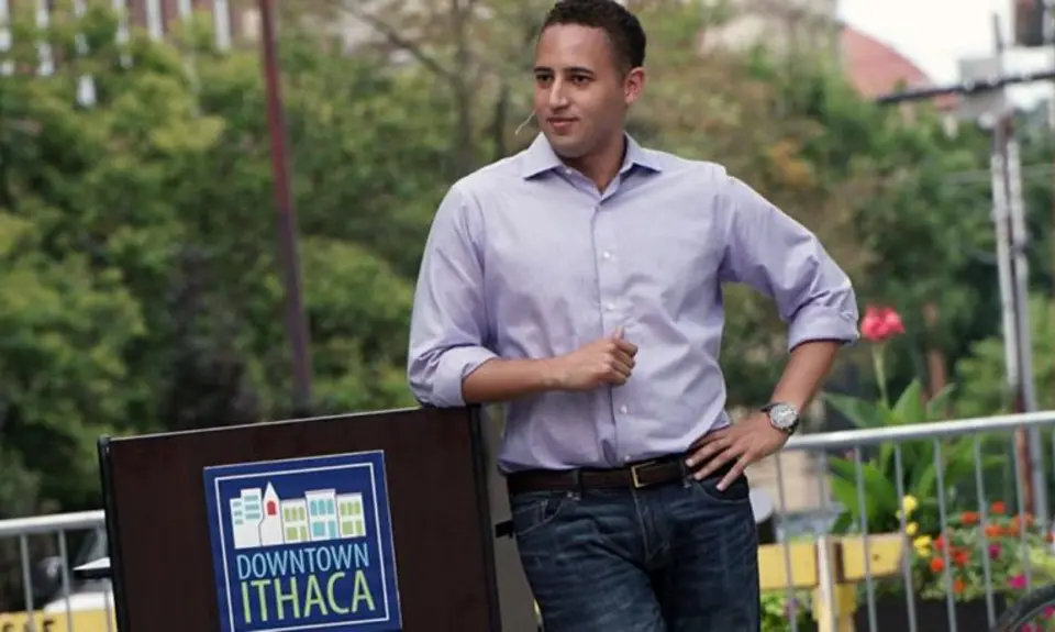 Svante Myrick wearing a purple shirt and standing next to a podium which says "Downtown Ithaca."