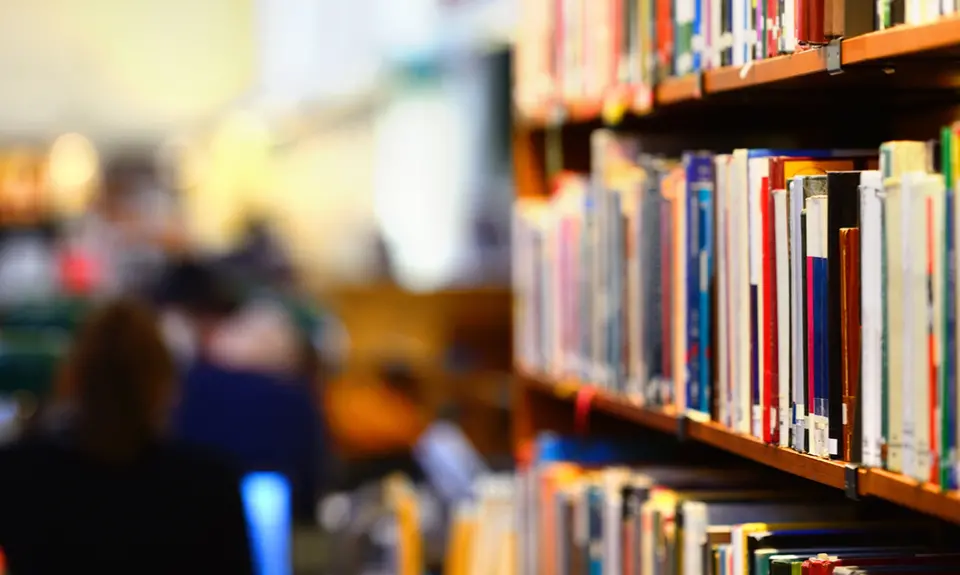 A full bookshelf in the foreground and people in a library in the background.