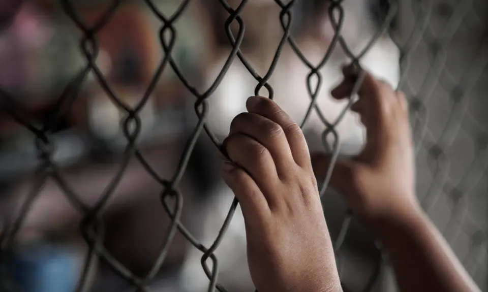 Two hands grip a chain link fence while other people are out of focus in the background.