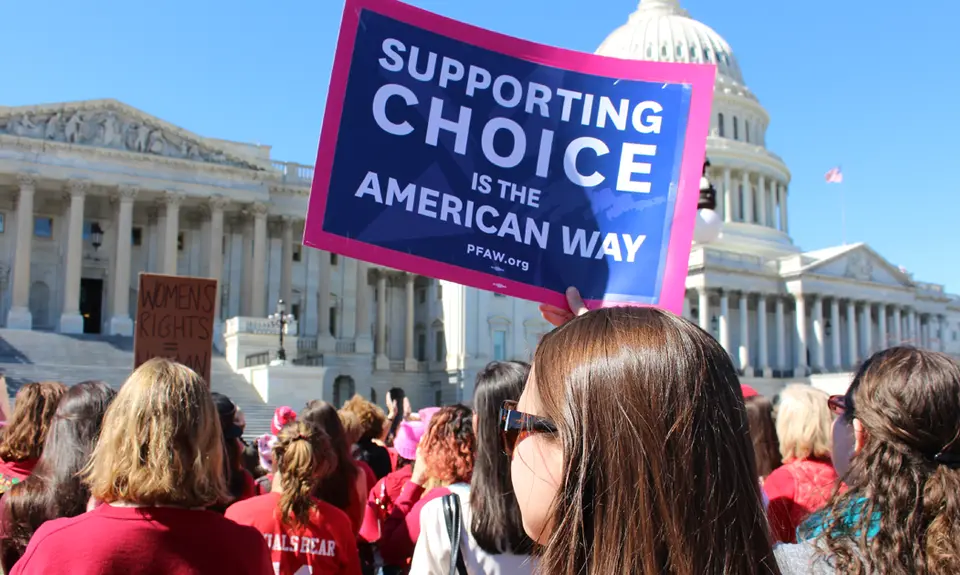 A group of people stand in front of the Capitol Building; one woman holds a sign that says "Supporting Choice is the American Way"
