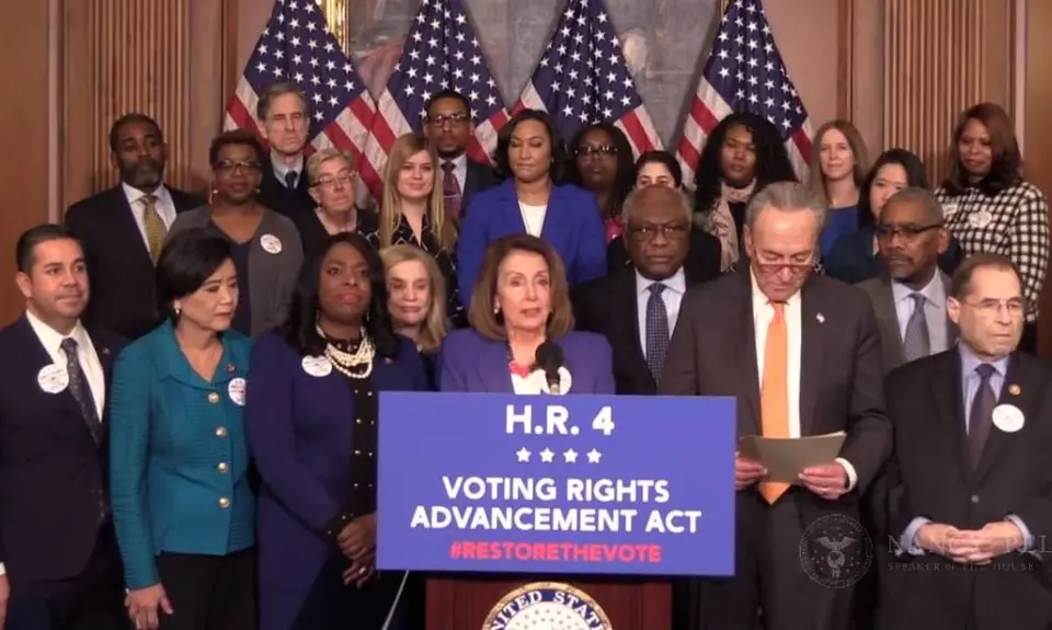 Nancy Pelosi stands at a podium with a group of people behind her while she talks about the Voting Rights Advancement Act