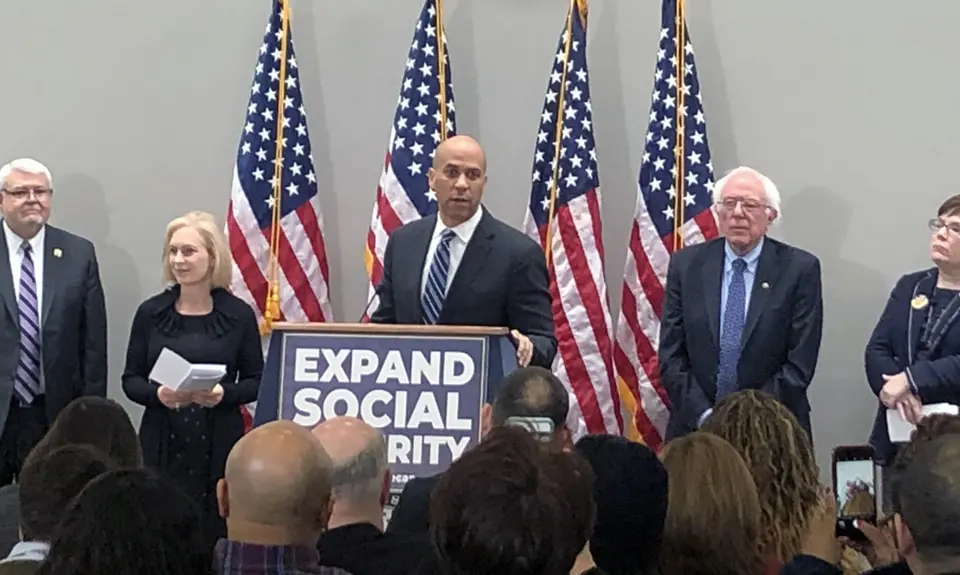 A group of lawmakers including Cory Booker and Bernie Sanders stand behind a podium with a sign that says "Expand Social Security"