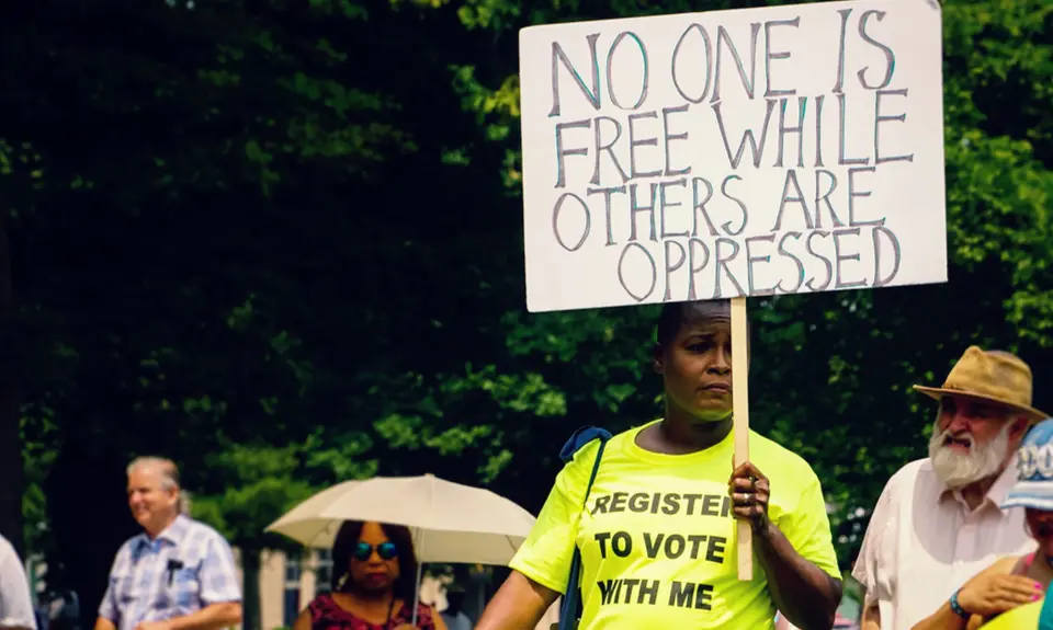 A person wearing a shirt that says "Register to Vote with Me" carries a sign that says "No one is free while others are oppressed."