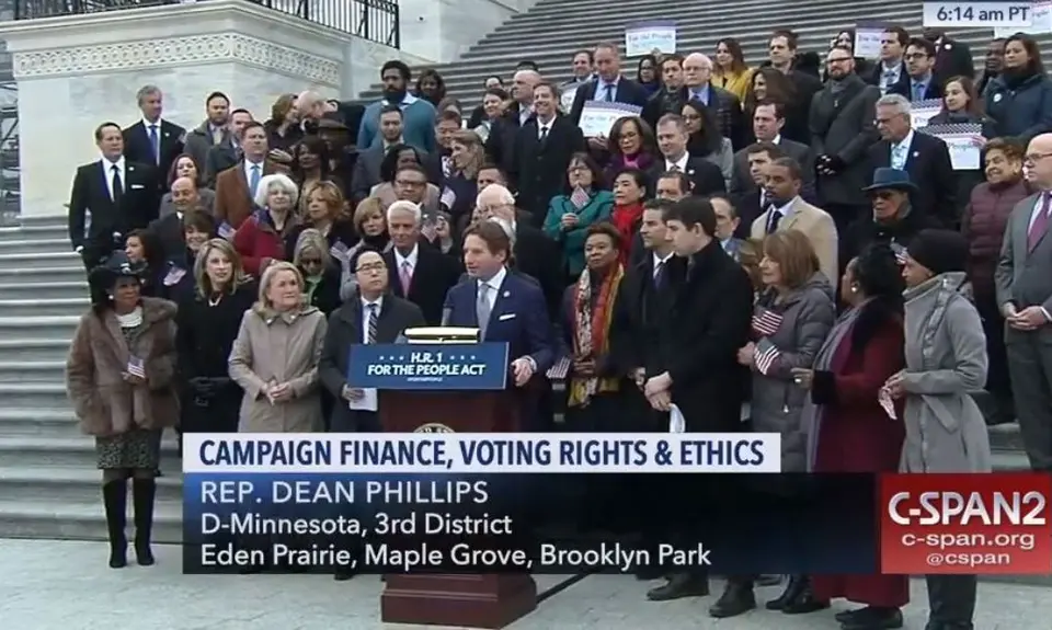 A screenshot from C-SPAN2 where a group of people stand behind Rep. Dean Phillips on a set of stairs as he talks about campaign finance, voting rights, and ethics.