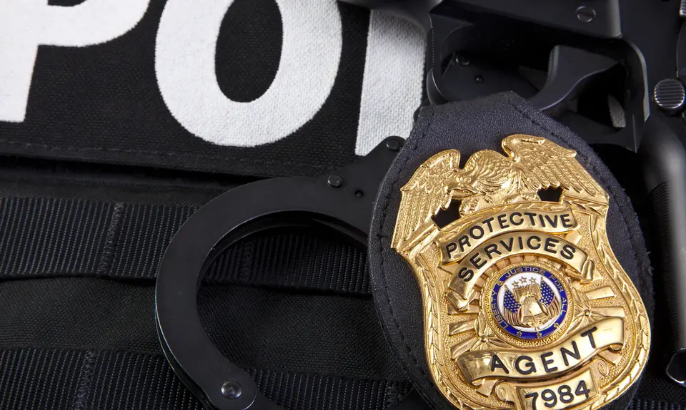 Closeup of a metal badge that says "Protective Services Agent" on a police vest.