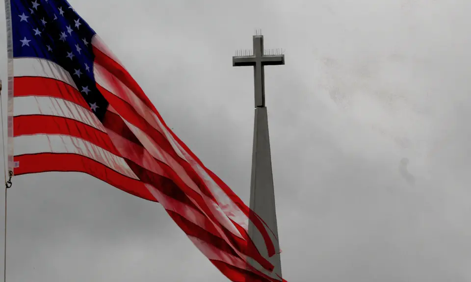 An American flag waves in front of the steeple of a church that is topped with a cross