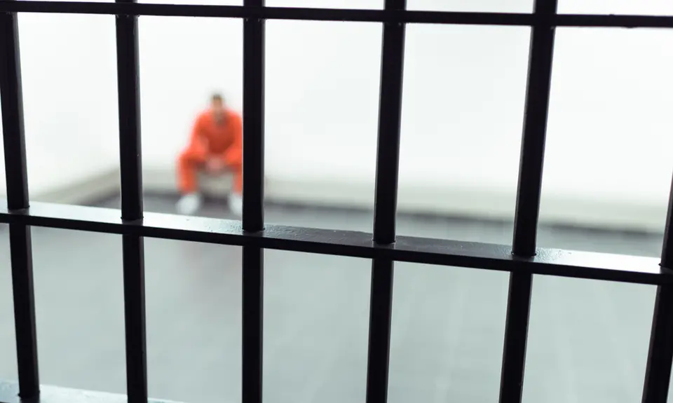 An image of a prisoner in an orange jumpsuit shown seated in a corner behind iron bars