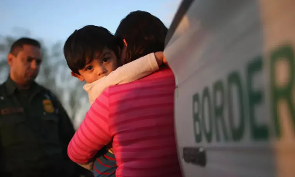 A young child clings to his mother near a border patrol truck and agent.