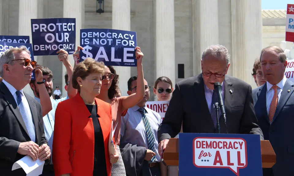 Chuck Schumer speaks at a podium while people around him hold signs saying "Protect our democracy" and "Democracy is not for sale."