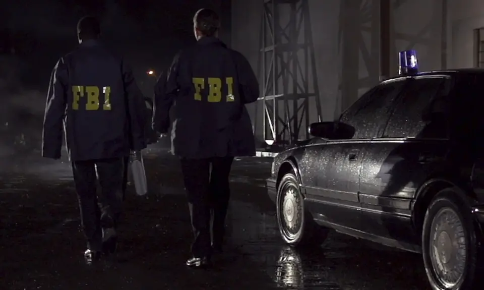 Two men in FBI jackets walk past a black car at night.