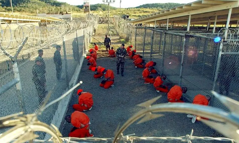 Detainees in orange jumpsuits sit on their knees and face a chain link fence at Camp X-Ray in Guantanamo Bay