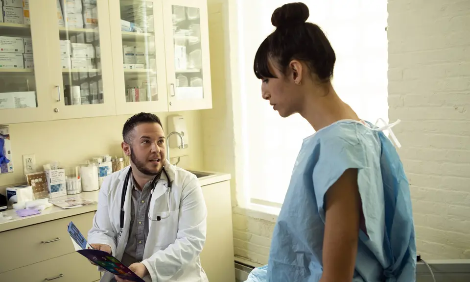 A transgender woman in a hospital gown speaking to her doctor, a