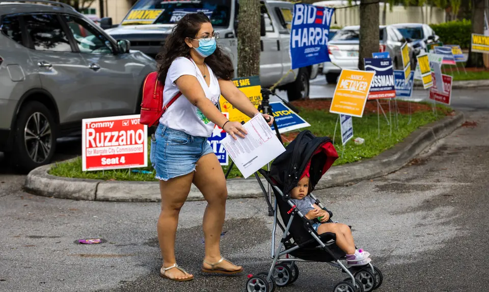 A Latina woman wearing a mask pushes her child in a stroller, with several political campaign signs behind her in the background.