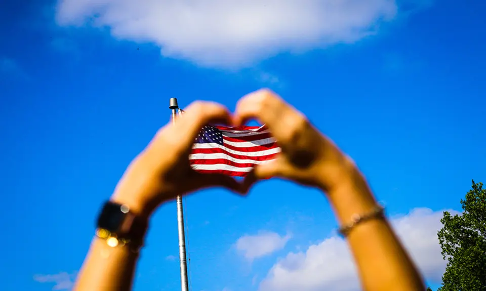 Two hands form a hard with their fingers in front of an American flag and blue sky.