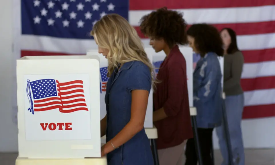 Four women cast ballots at voting booths with an American flag in the background.