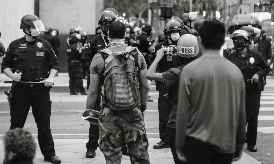 Two Black men and a reporter face a line of police in riot gear.