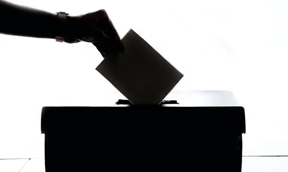 A silhouette of a hand dropping a ballot in a ballot box.