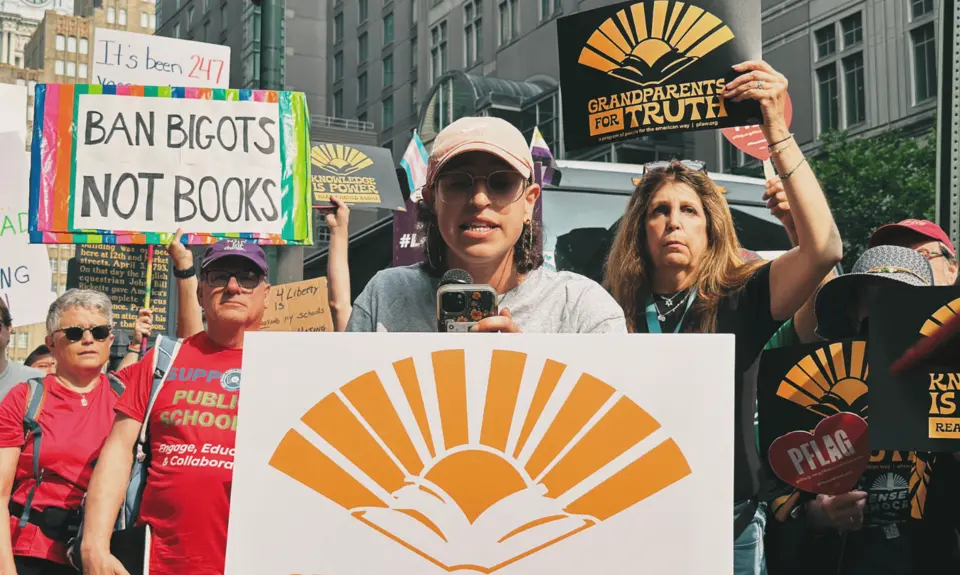 People at a rally. Older woman holds a black sign with yellow letters that reads "grandparents for truth"