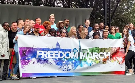 A couple dozen speakers and singers gather on stage behind a rainbow-colored "Freedom March" banner.