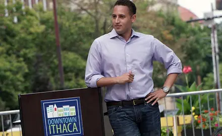 Svante Myrick wearing a purple shirt and standing next to a podium which says "Downtown Ithaca."