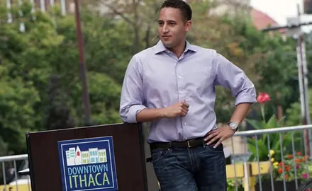 Svante Myrick wearing a purple shirt and standing next to a podium which says "Downtown Ithaca."