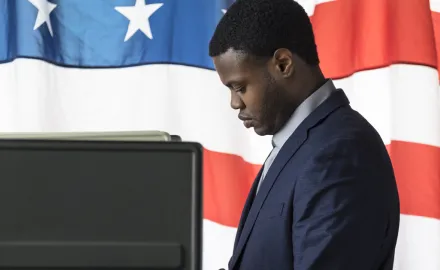 A Black man in a suit stands at a voting booth with an American flag in the background.