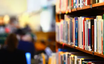 A full bookshelf in the foreground and people in a library in the background.