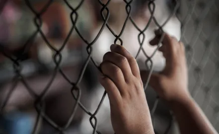 Two hands grip a chain link fence while other people are out of focus in the background.