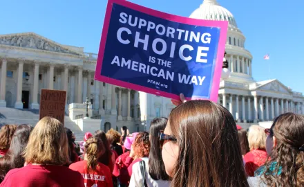 A group of people stand in front of the Capitol Building; one woman holds a sign that says "Supporting Choice is the American Way"