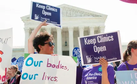 A group of people stand in front of the Supreme Court building with signs that say "Our bodies, our lives" and "Keep clinics open."