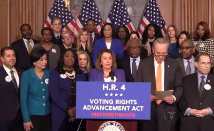 Nancy Pelosi stands at a podium with a group of people behind her while she talks about the Voting Rights Advancement Act