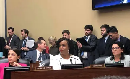 Rep. Ocasio-Cortez and Rep. Ayanna Pressley sit in a room during a hearing