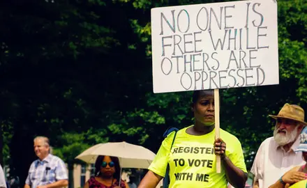 A person wearing a shirt that says "Register to Vote with Me" carries a sign that says "No one is free while others are oppressed."