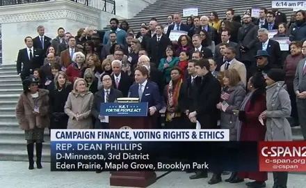 A screenshot from C-SPAN2 where a group of people stand behind Rep. Dean Phillips on a set of stairs as he talks about campaign finance, voting rights, and ethics.