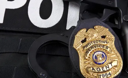 Closeup of a metal badge that says "Protective Services Agent" on a police vest.