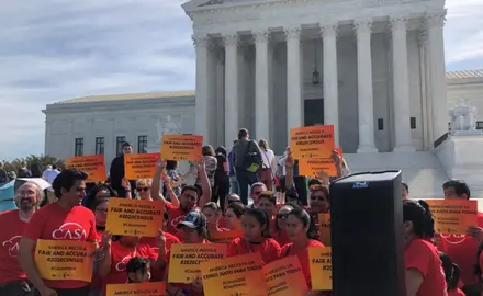 Activists stand outside the Supreme Court wearing red and carrying signs that read "America needs a fair and accurate #2020Census."