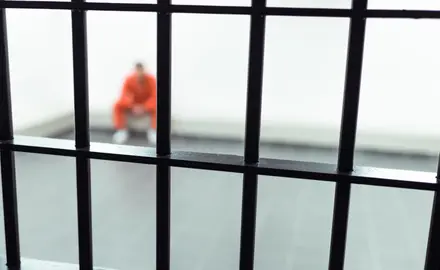 An image of a prisoner in an orange jumpsuit shown seated in a corner behind iron bars