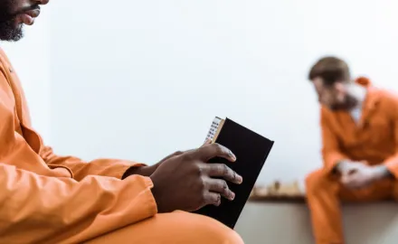 Two men in orange jumpsuits; the closest to the camera holds a book, the other is blurry and looks at a chess board in the background