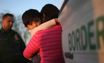 A young child clings to his mother near a border patrol truck and agent.