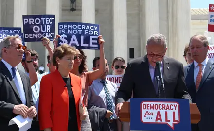 Chuck Schumer speaks at a podium while people around him hold signs saying "Protect our democracy" and "Democracy is not for sale."