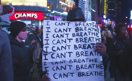 A man holds a sign in a crowded city; the sign says "I can't breathe. I can't breathe. I can't breathe."