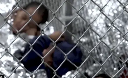 Children wrapped in emergency blankets laying behind a chain-link fence.