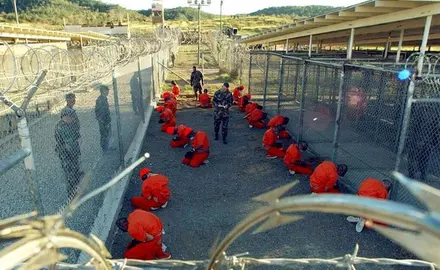 Detainees in orange jumpsuits sit on their knees and face a chain link fence at Camp X-Ray in Guantanamo Bay