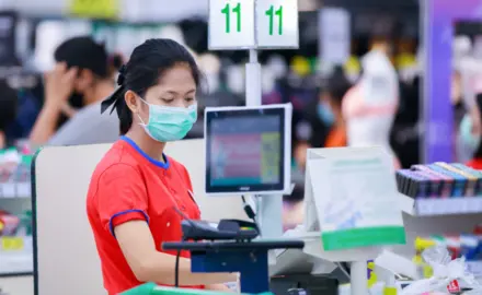 A young grocery store cashier wears a surgical mask while she works.